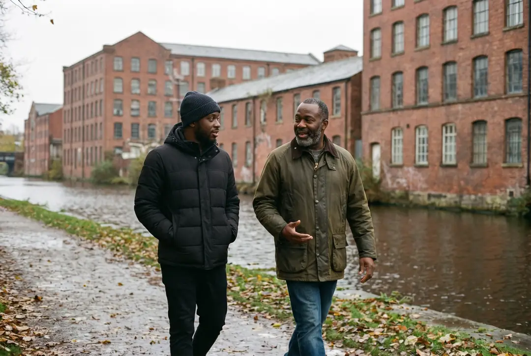 Two Black men, one younger in a beanie and one older with a grey beard, talk as they walk along a canal towpath past red-brick mills in Salford.
